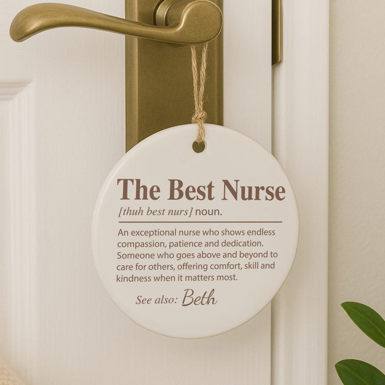 May include: A white, round door hanger with the text "The Best Nurse" in brown lettering. The hanger is suspended by a natural twine string from a gold-colored door handle. The text includes a definition of a nurse.