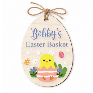 May include: Wooden Easter egg-shaped sign with the text "Bobby's Easter Basket" in blue script. The sign features a yellow chick hatching from a pink and white striped egg, surrounded by flowers and Easter eggs, and is topped with a twine bow.