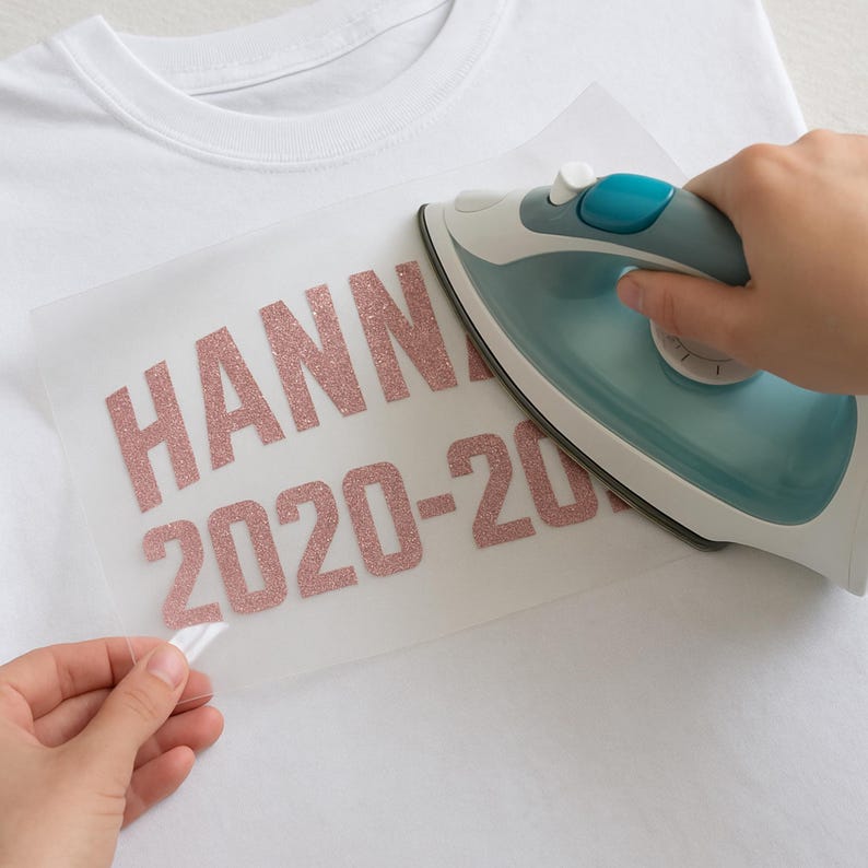 May include: A white t-shirt with a pink glitter iron-on design that says "HANNAH 2020-2021". A person is using an iron to press the design onto the shirt.