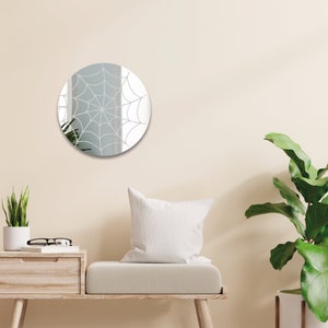 May include: A round mirror with a white spiderweb design. The mirror is hanging on a light beige wall above a light brown wooden bench with a white pillow.