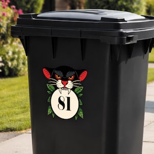 May include: Black trash bin with a decorative sticker featuring a black panther design. The panther has red ears, yellow eyes, and a white-toothed grin. A white circle with the number 81 is surrounded by green leaves.