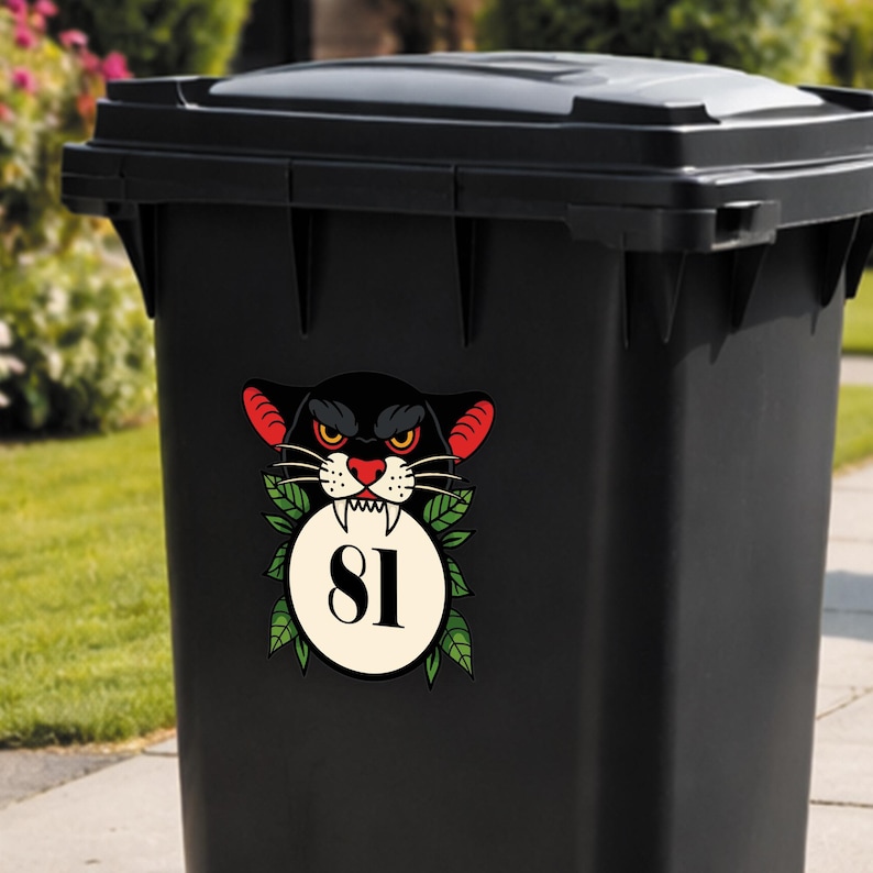 May include: Black trash bin with a decorative sticker featuring a black panther design. The panther has red ears, yellow eyes, and a white-toothed grin. A white circle with the number 81 is surrounded by green leaves.