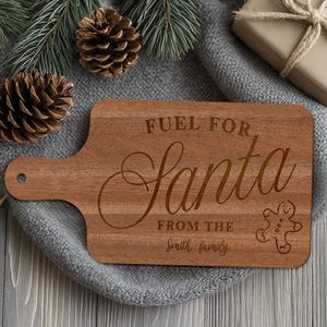 May include: A wooden cutting board with a handle, engraved with "FUEL FOR Santa" and "FROM THE Smith family", plus a gingerbread man. The board is brown and rests on a gray surface, with pinecones and greenery in the background.
