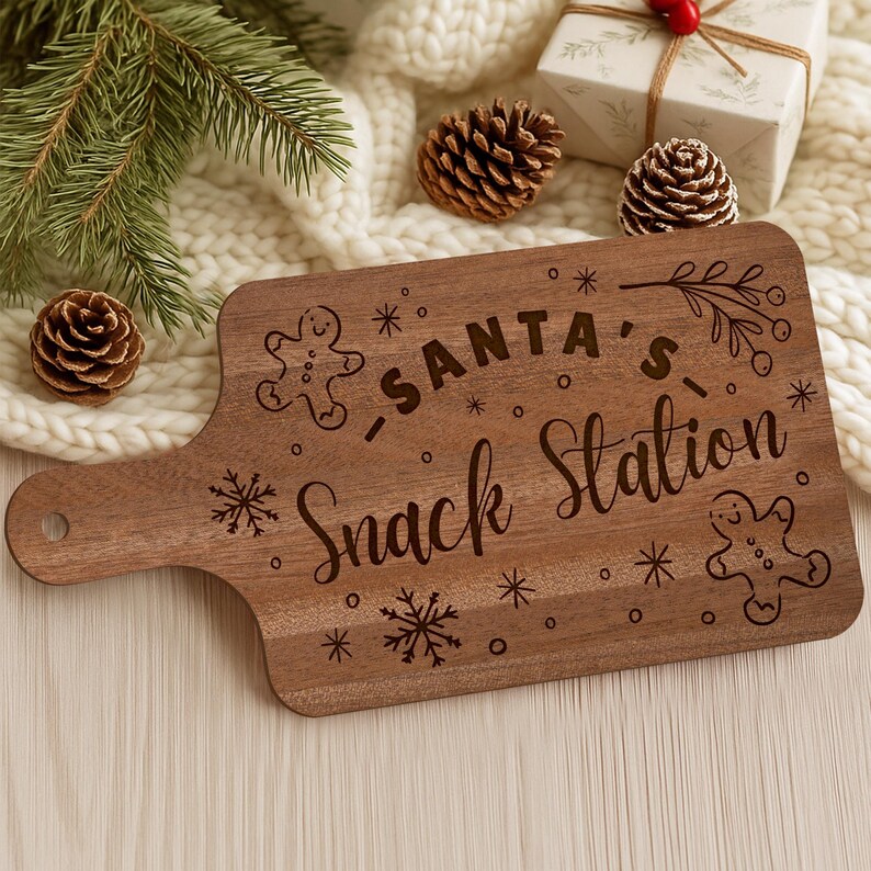 May include: A wooden cutting board with the words "Santa's Snack Station" engraved on it. The board features gingerbread men and snowflake designs. The board has a handle with a hole for hanging. The background includes pine cones, a gift, and evergreen branches.