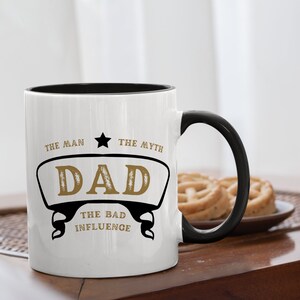 May include: White ceramic mug with a black rim and handle. The mug has a gold design that says "The Man, The Myth, Dad, The Bad Influence".