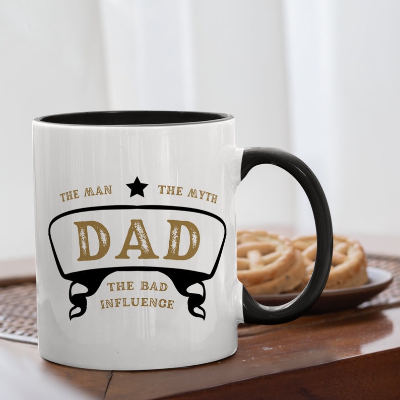 May include: White ceramic mug with a black rim and handle. The mug has a gold design that says "The Man, The Myth, Dad, The Bad Influence".