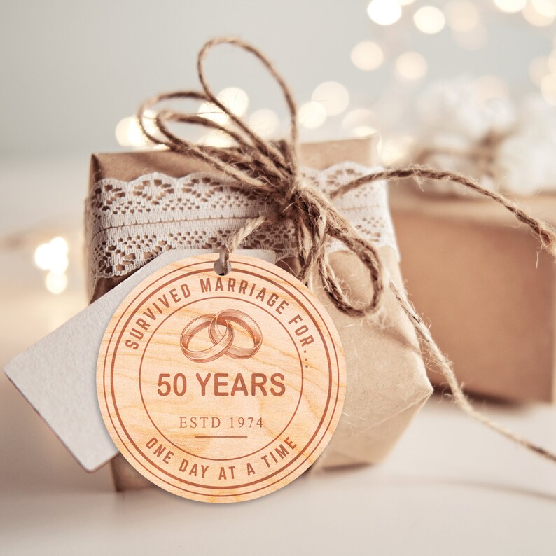 May include: A round wooden tag with the text "Survived Marriage For... 50 Years Estd 1974 One Day At A Time" and a graphic of two interlocking rings.