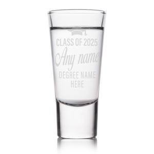 May include: Clear shot glass with etched text: "CLASS OF 2025", "Any name", and "DEGREE NAME HERE". A graduation cap graphic is above the text. The glass is cylindrical with a thick base and is filled with a clear liquid.
