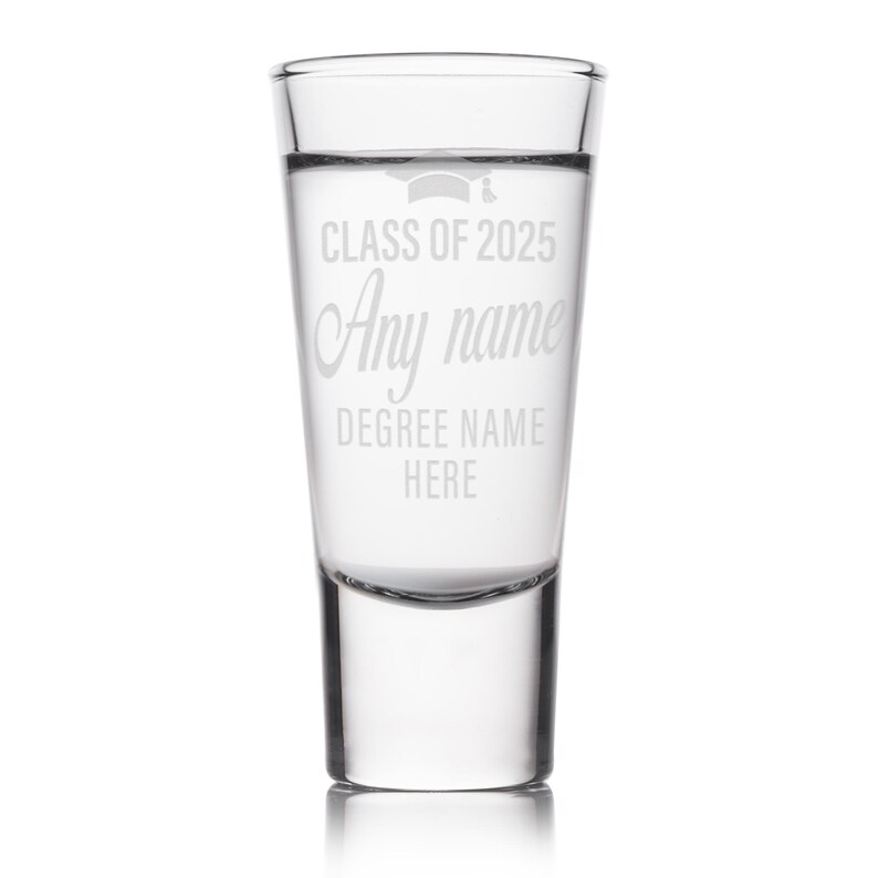May include: Clear shot glass with etched text: "CLASS OF 2025", "Any name", and "DEGREE NAME HERE". A graduation cap graphic is above the text. The glass is cylindrical with a thick base and is filled with a clear liquid.
