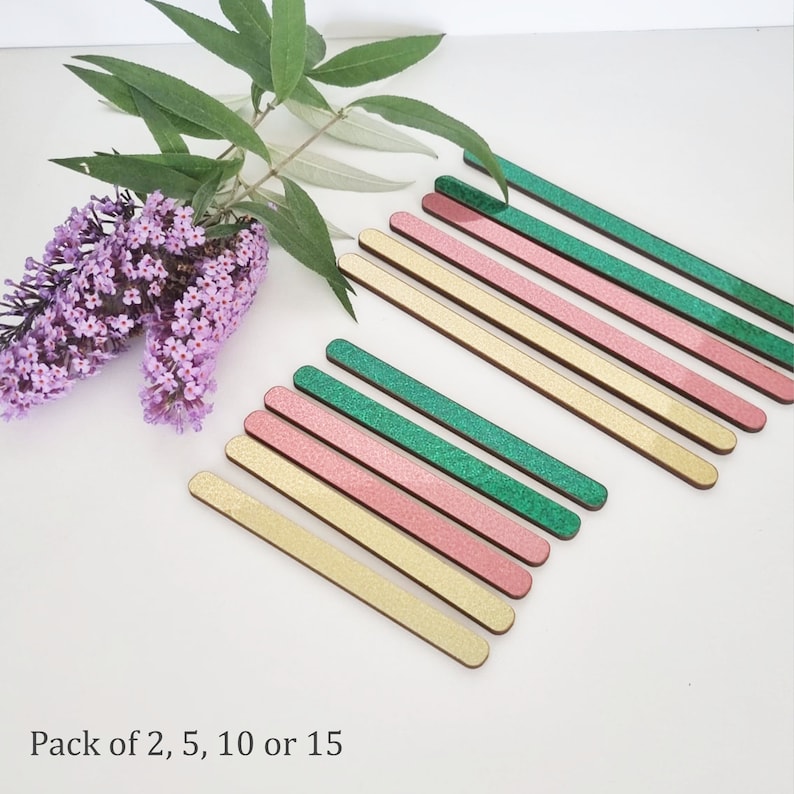 May include: A set of 15 wooden craft sticks with glitter. The sticks are in three colors: green, pink, and gold. Pack of 2, 5, 10 or 15.