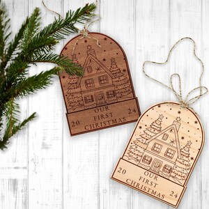 May include: Two wooden Christmas ornaments with a house and snow scene design. The ornaments are engraved with "Our First Christmas" and the numbers 20 and 24.