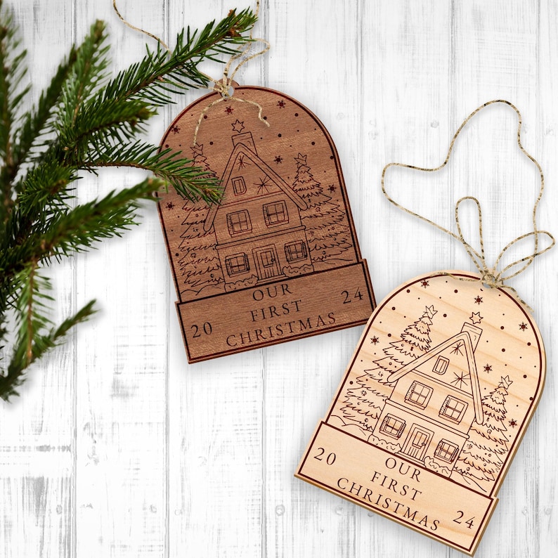 May include: Two wooden Christmas ornaments with a house and snow scene design. The ornaments are engraved with "Our First Christmas" and the numbers 20 and 24.