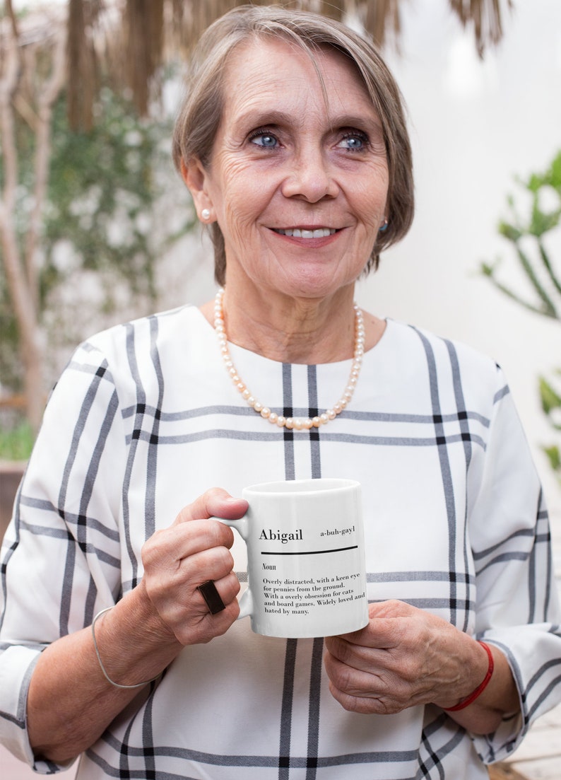 May include: White ceramic mug with a black and white definition of the name Abigail. The mug is being held by a person wearing a white and black plaid shirt.