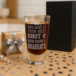 May include: A clear shot glass filled with a dark liquid, featuring the etched text "YOU GAVE IT YOUR BEST SHOT & NOW YOU'RE A GRADUATE." A wrapped gift with a silver bow and a black box are in the background. The table is decorated with star-shaped confetti.