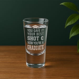 May include: A clear shot glass filled with liquid, featuring the etched text "YOU GAVE IT YOUR BEST SHOT & NOW YOU'RE A GRADUATE." The glass has a graduation cap graphic above the text. The glass is on a wooden surface.