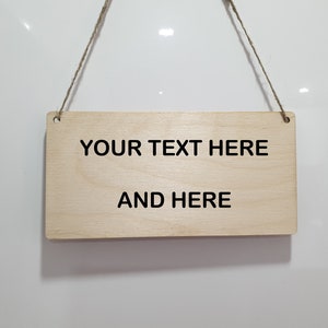 Puede incluir: Un letrero rectangular de madera clara con el texto "YOUR TEXT HERE" en la l&iacute;nea superior y "AND HERE" en la inferior, en fuente negra. El letrero est&aacute; suspendido por una cuerda de hilo natural.