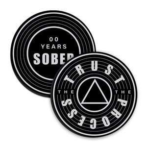 May include: Two black round coasters with white text. The first coaster has the text "00 YEARS SOBER". The second coaster has the text "TRUST THE PROCESS" with a black triangle in the centre.