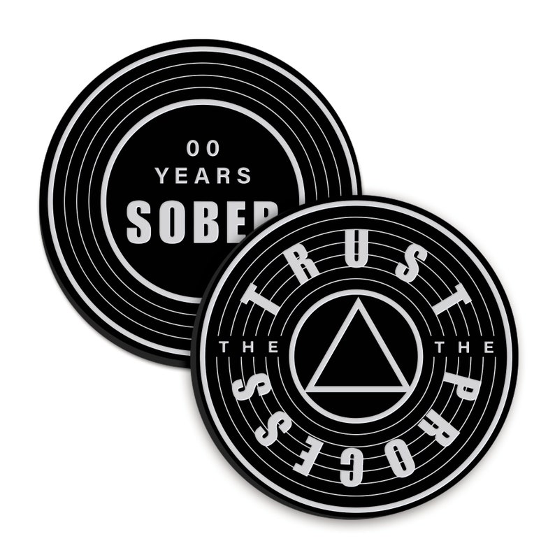 May include: Two black round coasters with white text. The first coaster has the text "00 YEARS SOBER". The second coaster has the text "TRUST THE PROCESS" with a black triangle in the centre.