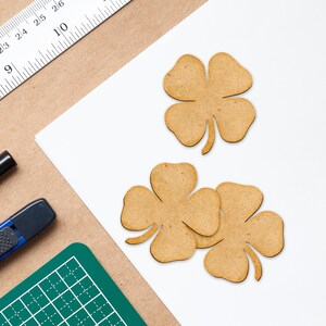 May include: Three wooden four-leaf clover cutouts in a light brown color, arranged on a white surface. A ruler, a green cutting mat, and a black marker are visible in the background. The clovers are likely for crafting or decoration.