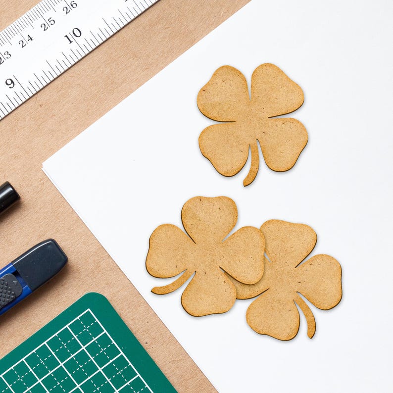 May include: Three wooden four-leaf clover cutouts in a light brown color, arranged on a white surface. A ruler, a green cutting mat, and a black marker are visible in the background. The clovers are likely for crafting or decoration.