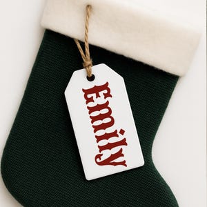 May include: A dark green Christmas stocking with a white cuff and a white tag. The tag is attached with twine and features the name "Emily" in red, decorative lettering. The stocking is made of a textured fabric.
