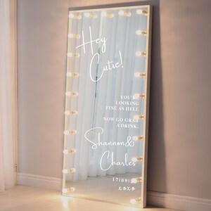May include: A white vanity mirror with a gold frame and light bulbs around the perimeter. The mirror has a white inscription that reads "Hey Cutie! You're looking fine as hell. Now go grab a drink. Shannon & Charles 171091 xoxo"