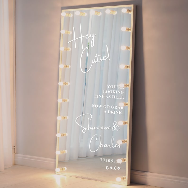 May include: A white vanity mirror with a gold frame and light bulbs around the perimeter. The mirror has a white inscription that reads "Hey Cutie! You're looking fine as hell. Now go grab a drink. Shannon & Charles 171091 xoxo"