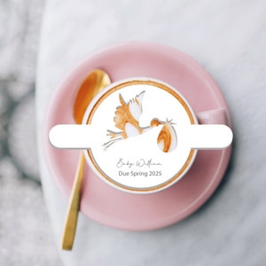 May include: A pink saucer with a white coffee cup. The coffee cup has a white stencil design of a stork carrying a baby. The text "Baby William Due Spring 2025" is written below the design.