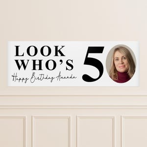 May include: A white banner with the text "LOOK WHO'S 50" and "Happy Birthday Amanda" in black. A photo of a woman is on the right. The banner is likely for a 50th birthday celebration.