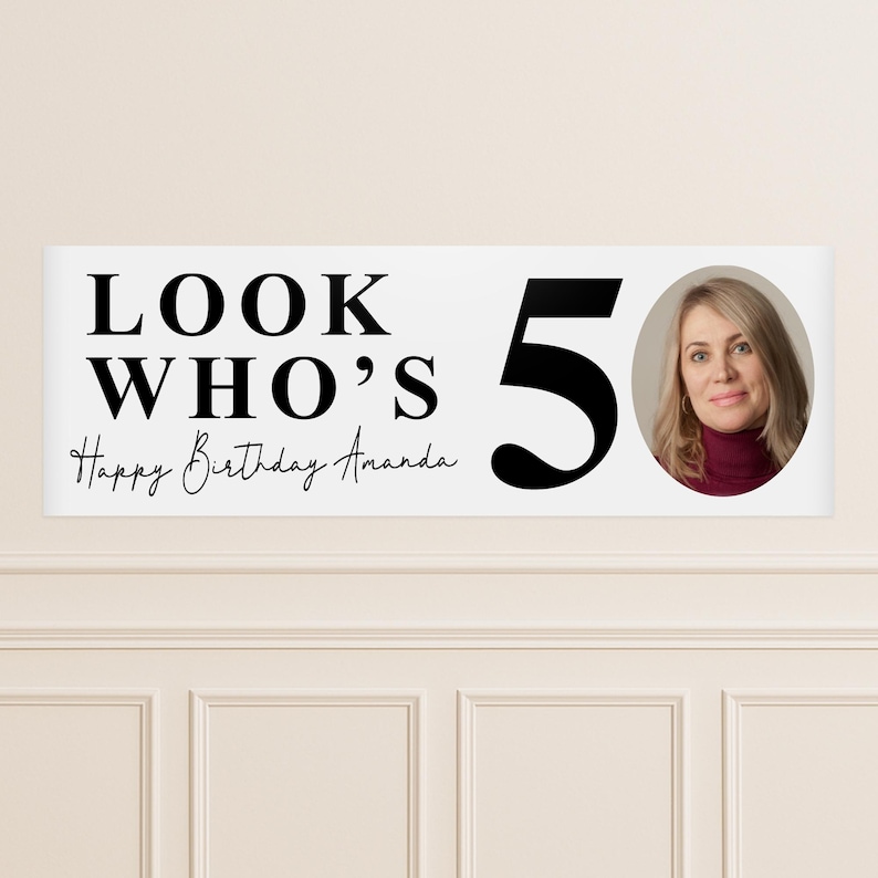 May include: A white banner with the text "LOOK WHO'S 50" and "Happy Birthday Amanda" in black. A photo of a woman is on the right. The banner is likely for a 50th birthday celebration.