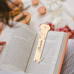 May include: A wooden bookmark shaped like a dog with a cute face and the name "Jamie" engraved on it. The bookmark is light brown and has a natural wood finish.