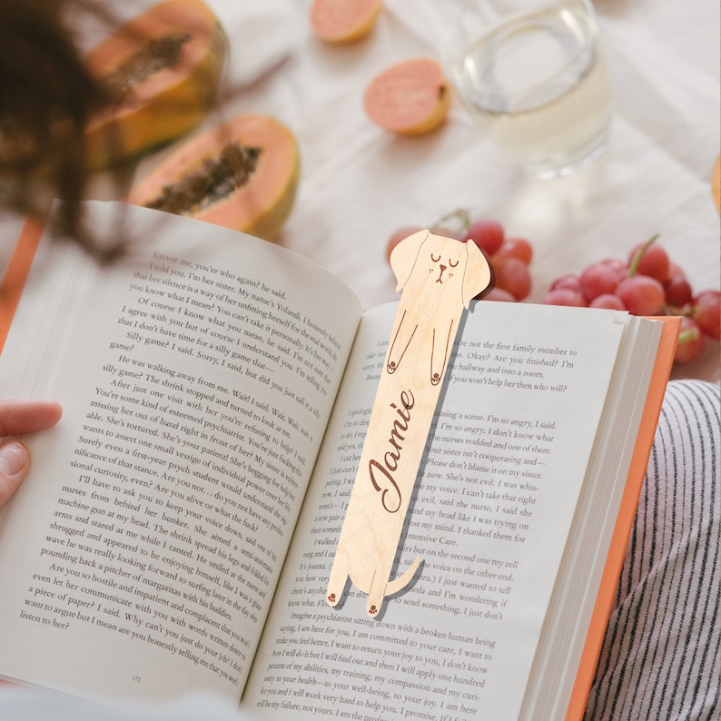 May include: A wooden bookmark shaped like a dog with a cute face and the name "Jamie" engraved on it. The bookmark is light brown and has a natural wood finish.