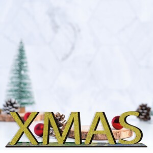 K&ouml;nnte beinhalten: Ein goldener Glitzer-&bdquo;XMAS&ldquo;-Schriftzug mit roten Kugeln auf einem Holzsockel. Das Schild steht auf einer wei&szlig;en Marmorfl&auml;che mit einem verschwommenen gr&uuml;nen Weihnachtsbaum im Hintergrund.