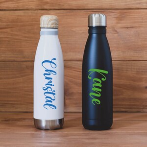 May include: Two stainless steel water bottles with personalised names. The bottle on the left is white with the name "Christal" in blue lettering. The bottle on the right is black with the name "Kaume" in green lettering.