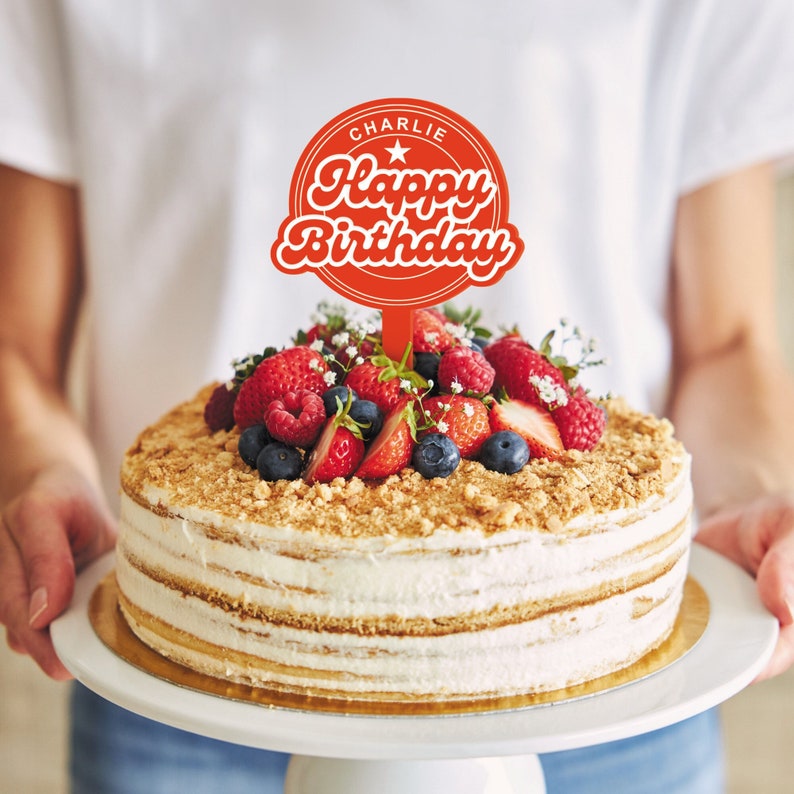 May include: A red cake topper with the text "Happy Birthday" and the name "Charlie" in a circle. The topper is on a cake decorated with fresh berries and white frosting.