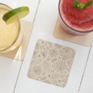 May include: Two drinks on coasters with a leaf pattern. The drink on the left is a yellow beverage with a lime wedge. The drink on the right is a red smoothie with a sprig of mint.