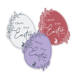 May include: Three oval-shaped stickers with a floral design and the text "Olivia's First Easter 2024". The stickers are in white, pink, and purple.