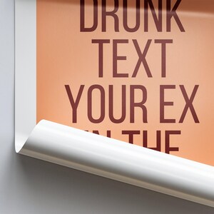 Op de afbeelding: Een close-up van een poster met de tekst "DRUNK TEXT YOUR EX IN THE" gedrukt in donkerrood op een perzikkleurige achtergrond. De poster is gedeeltelijk opgerold, waardoor een witte rand zichtbaar is.