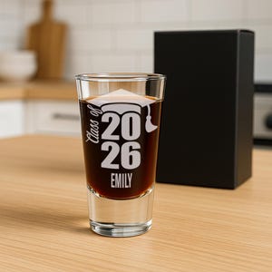 May include: A clear shot glass filled with a dark liquid, featuring the text "Class of 2026" and a graduation cap graphic. The name "Emily" is printed below the year. A black gift box is in the background.