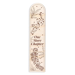 Op de afbeelding: Houten bladwijzer in lichtbruin met gravures. Het ontwerp toont illustraties van luipaarden en bladeren, met de tekst "One More Chapter". Het ontwerp omvat een springende luipaard en een met een welp. De vorm is rechthoekig met een afgeronde bovenkant.