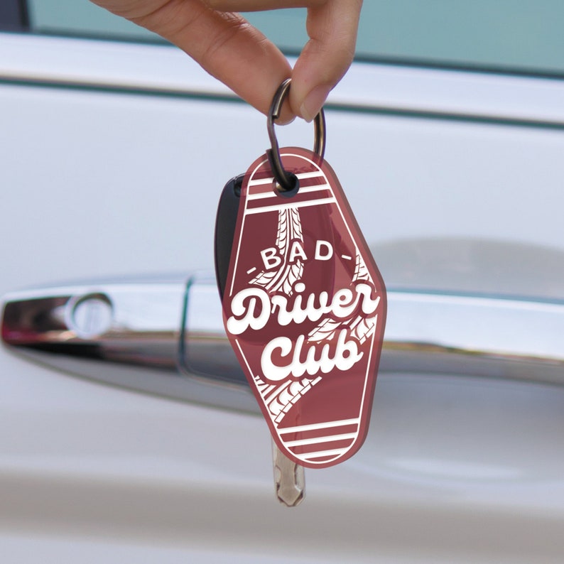 May include: A red motel key chain with the text "Bad Driver Club" in white lettering. The key chain is attached to a silver key ring.