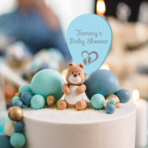 May include: A blue and white cake topper with a teddy bear and a balloon that says "Tammy's Baby Shower". The balloon has a heart with footprints on it. The cake is decorated with blue and gold balls.