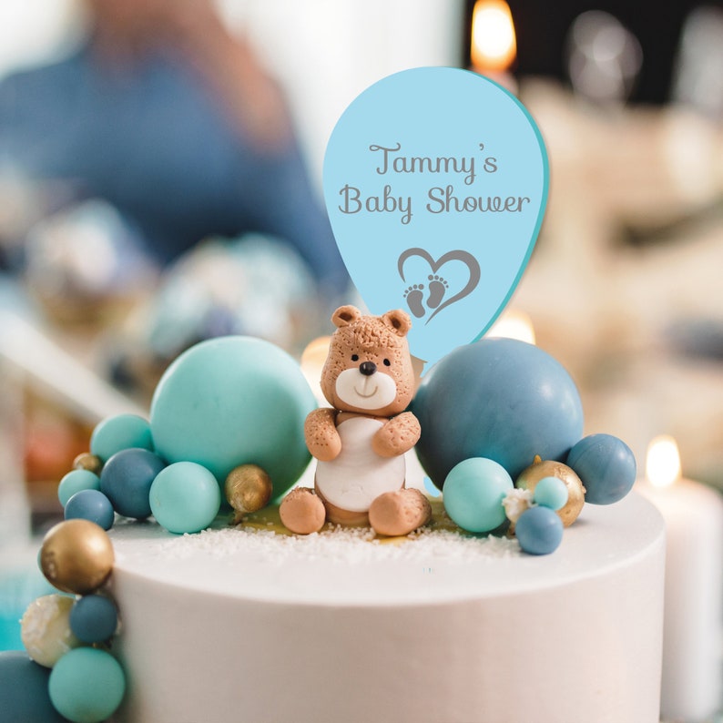 May include: A blue and white cake topper with a teddy bear and a balloon that says "Tammy's Baby Shower". The balloon has a heart with footprints on it. The cake is decorated with blue and gold balls.