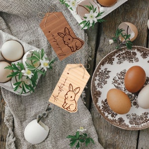 May include: Two wooden gift tags with bunny illustrations and handwritten names "Anastasia" and "Nicholas". The tags are on a burlap background with eggs and flowers.