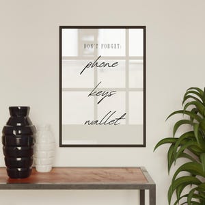 May include: A black framed print with the text "Don't forget: phone keys wallet" on a white background. The print is hanging on a white wall above a wooden shelf with two black and white vases.