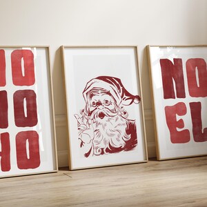 May include: Three framed Christmas prints. The center print features a red illustration of Santa Claus. The other two prints have the words "HO HO" and "NO EL" in red block letters. The frames are a light brown color.