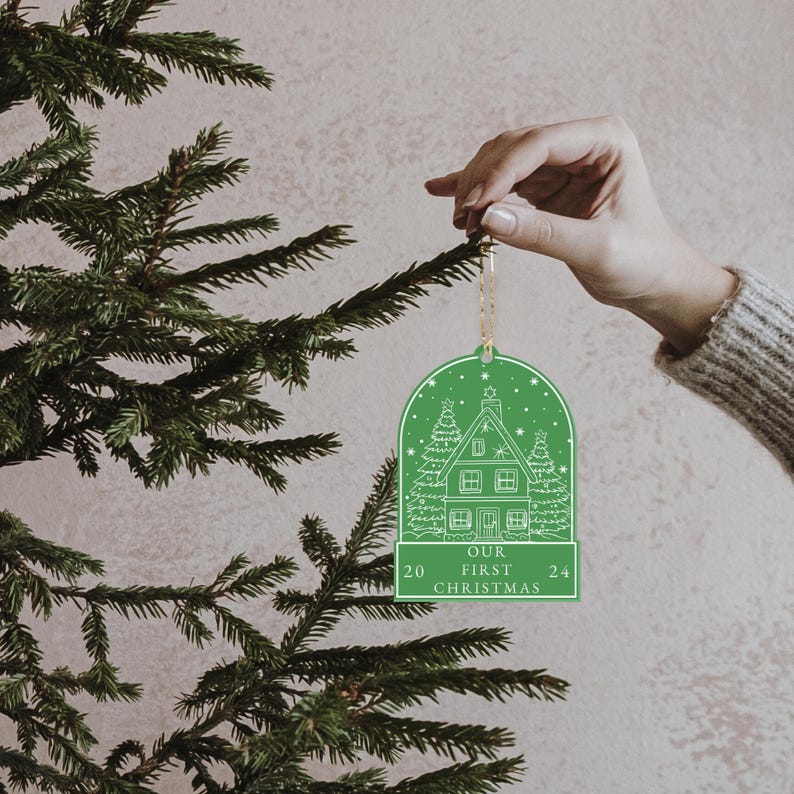 May include: Green Christmas ornament with a white outline of a house and trees. The ornament says "Our First Christmas 20 24".