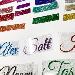 K&ouml;nnte beinhalten: Nahaufnahme von mehreren Bl&auml;ttern Glitzerpapier in verschiedenen Farben, darunter Pink, Lila, Gr&uuml;n und Gold. Das Glitzerpapier ist in Reihen auf einer wei&szlig;en Oberfl&auml;che angeordnet. Es gibt auch mehrere Bl&auml;tter aus klarem Vinyl mit Namen, die in Glitzer geschrieben sind, darunter "Alex", "Salt" und "Tasha".