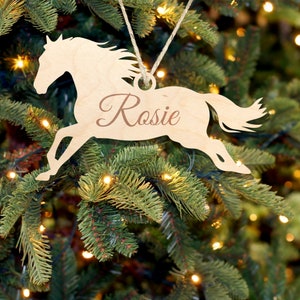 May include: Wooden horse ornament with the name "Rosie" written on it. The ornament is hanging on a Christmas tree with lights.