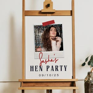 May include: A wooden easel displays a sign with a photo of a woman blowing a kiss. The sign reads "Sasha's HEN PARTY" with the date "09/10/25" below. A red piece of tape secures the photo.